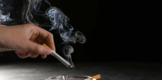 A hand holding a cigarette above an ashtray filled with used cigarettes and smoke rising