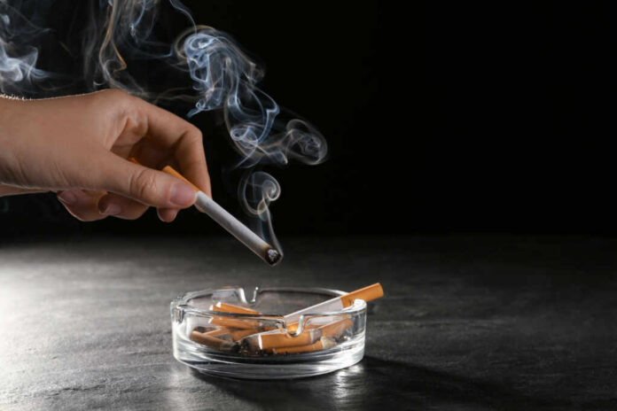 shutterstock_2127942221.jpg A hand holding a cigarette above an ashtray filled with used cigarettes and smoke rising