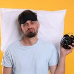 Man wearing a sleep mask holding an alarm clock with a frustrated expression