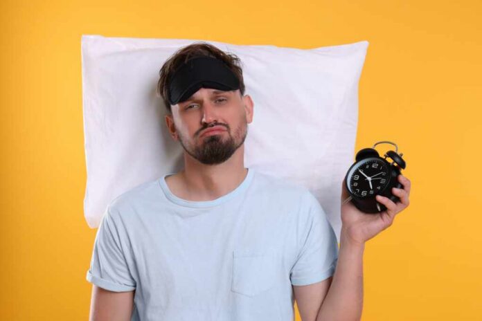 Man wearing a sleep mask holding an alarm clock with a frustrated expression