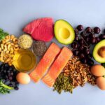 An assortment of healthy foods including fish, nuts, fruits, and vegetables arranged on a light background