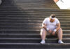 An exhausted man in sportswear sitting on outdoor stairs, looking down.