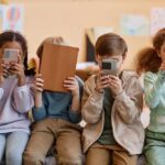 Four children sitting together, each engaged with their electronic devices
