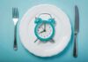 Fasting Earlier or Later in Day for Weight Loss? Alarm clock on a white plate with fork and knife