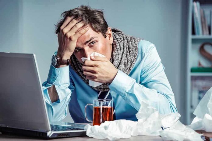 shutterstock_1112983769 Sick man at desk blowing his nose