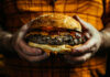 A person holding a large cheeseburger with lettuce, tomato, and condiments