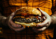 A person holding a large cheeseburger with lettuce, tomato, and condiments
