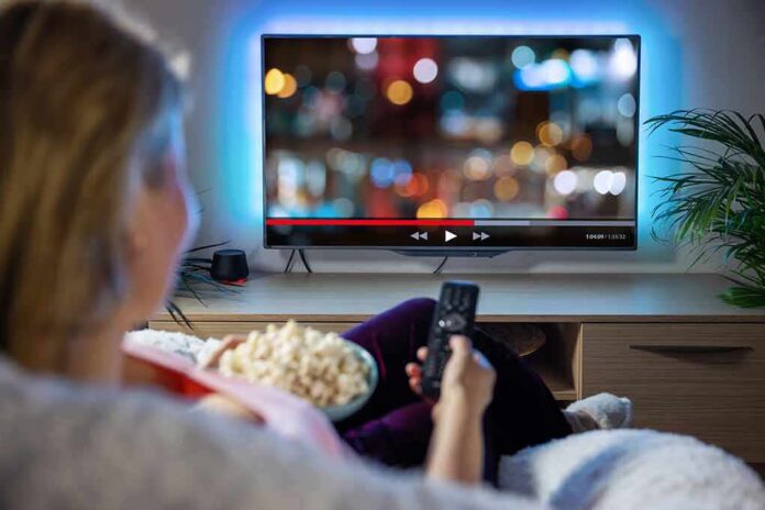 c0e4d3fd-3b50-4cd9-b02f-4e9933d13d92_shutterstock_1966720738 Person sitting on a couch watching a movie on a TV with popcorn in hand