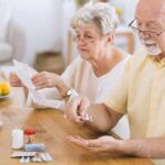 Older man taking pills at a table while woman reads instructions