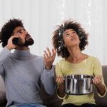 Couple on couch reacting to ceiling leak as woman holds pot to catch water