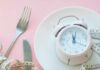 Aging Bodies Blindsided By Protein Timing Pink alarm clock on a white plate with fork, knife, and measuring tape