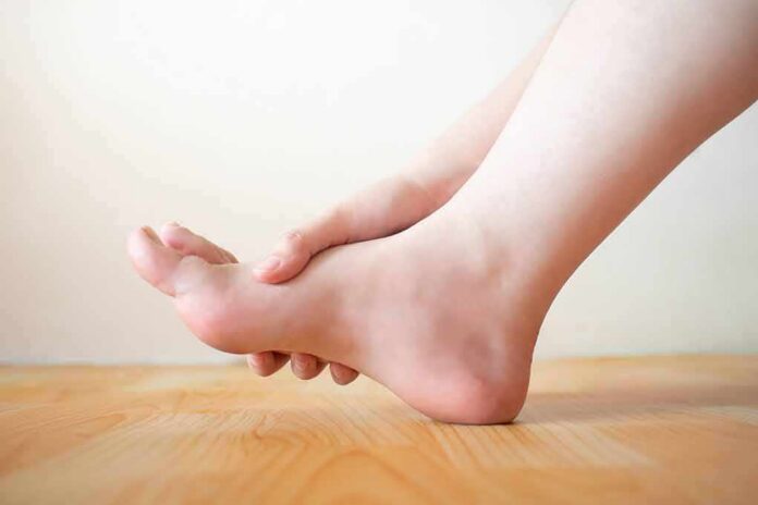 shutterstock_1186138735 Hand holding the arch of a bare foot on a wooden floor