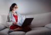 Person wearing orange glasses using a laptop on a white couch