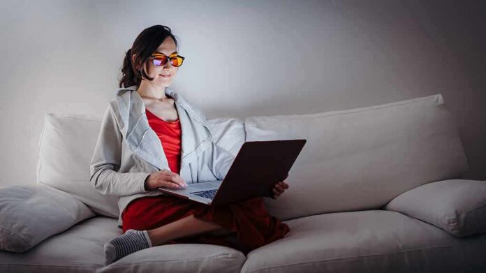shutterstock_1081014089 Person wearing orange glasses using a laptop on a white couch