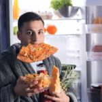 man in robe eating pizza at open refrigerator