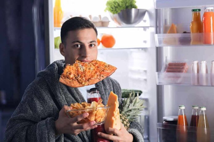 man in robe eating pizza at open refrigerator