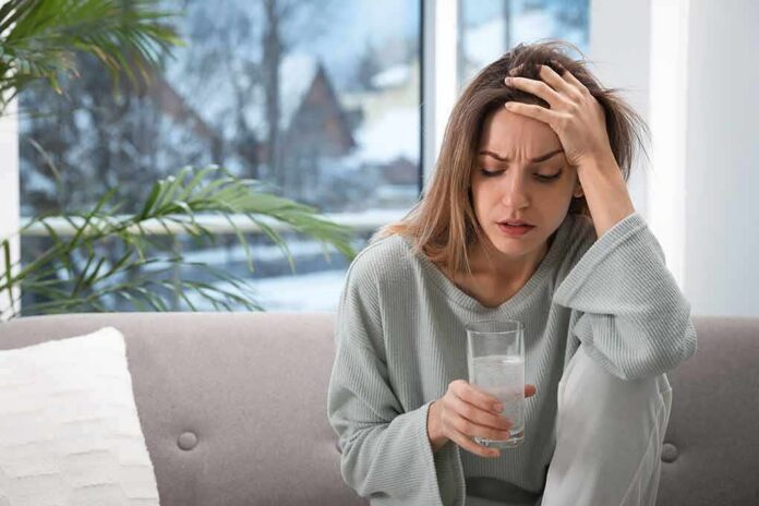 shutterstock_1912160086 Woman holding a glass of water with hand on forehead