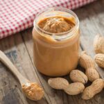 Jar of peanut butter with spoon and peanuts on rustic table